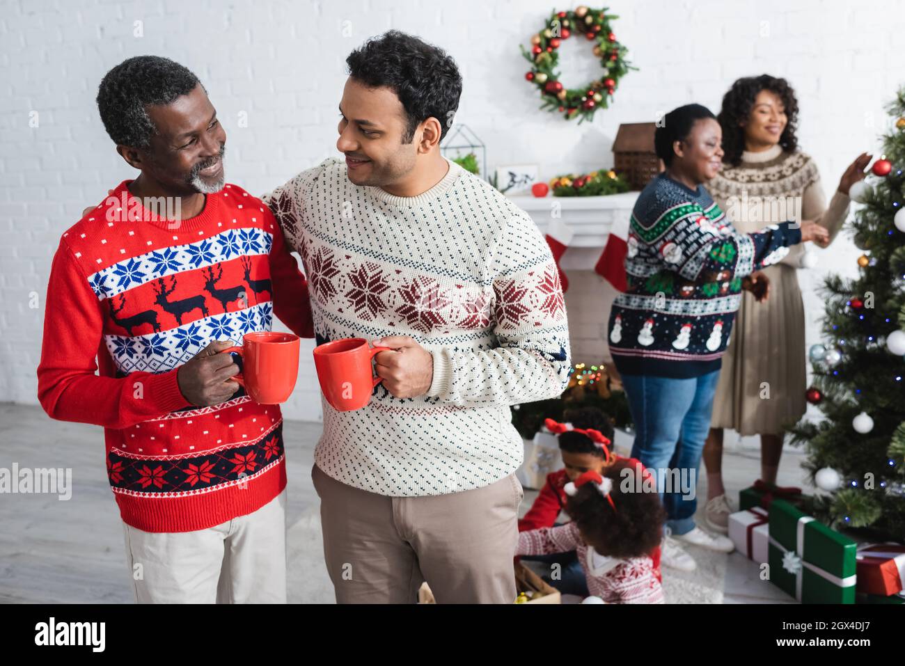 happy african american men with cups talking near blurred women and kids decorating christmas tree Stock Photo