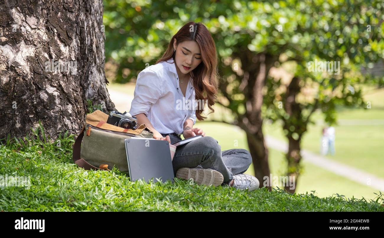 Image of joyful woman with diary book writing note while sitting on grass in park. Stock Photo