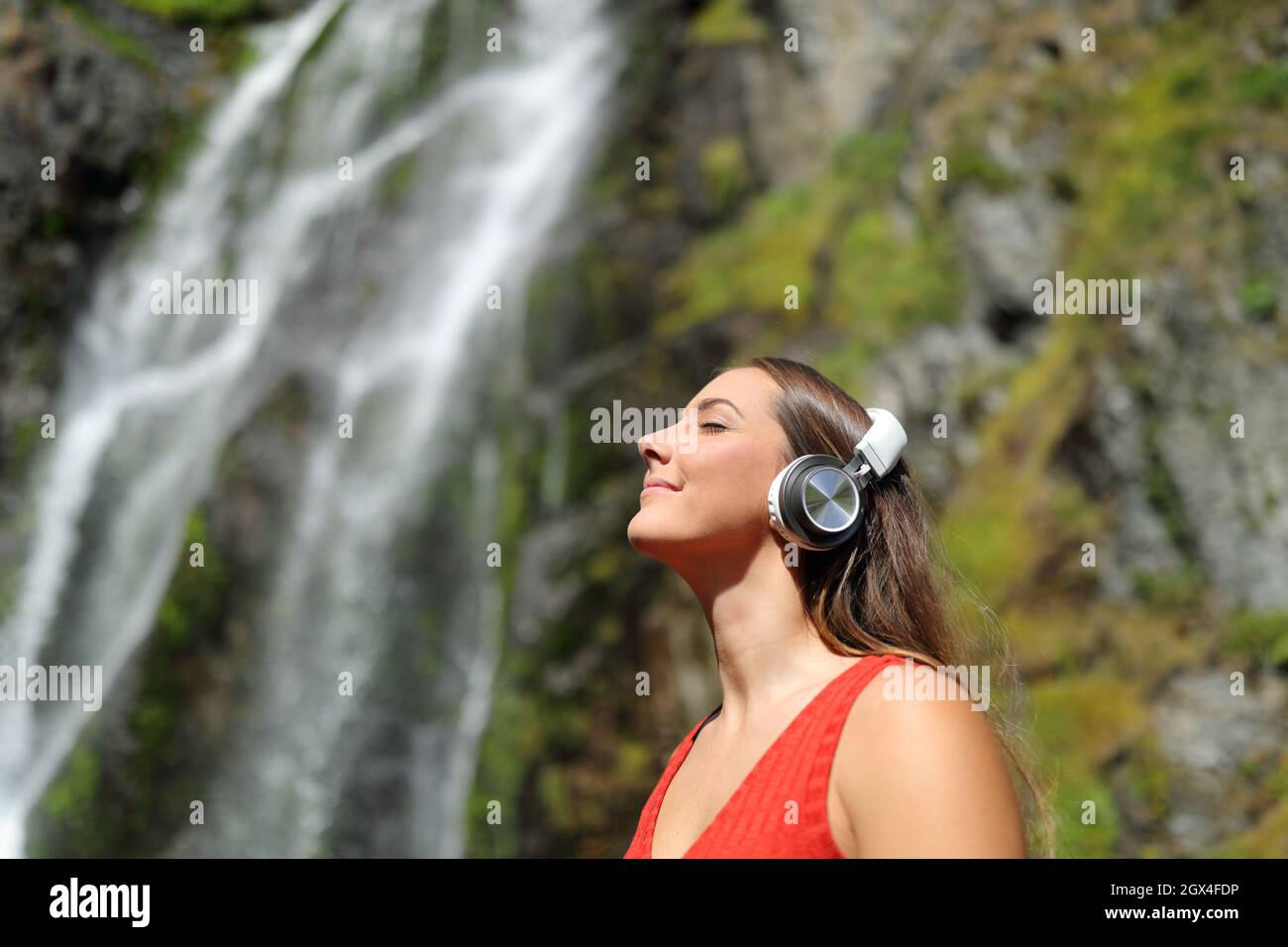 Happy woman in red listening to music wearing headphones breathing fresh air in a waterfall Stock Photo