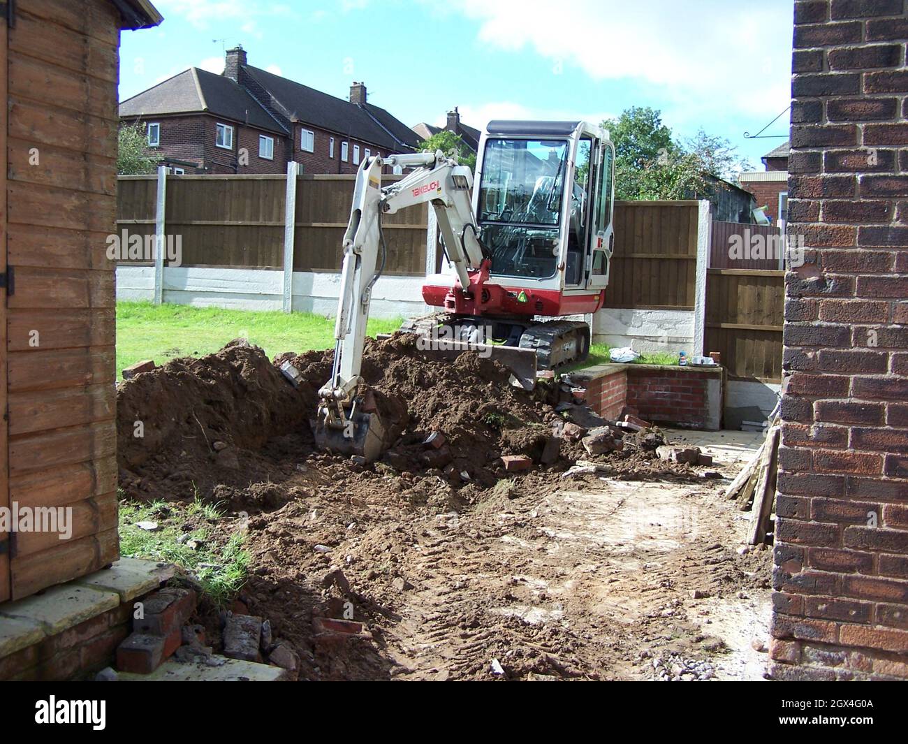 Workman using a mini digger to excavate a hole for extension footings in a garden lawn with green grass Stock Photo