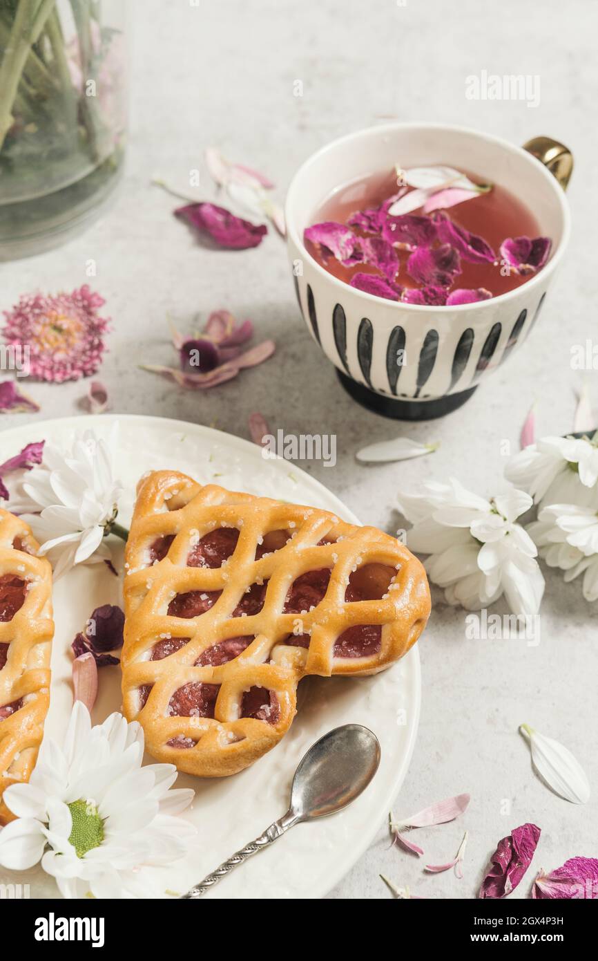 Close up of heart cake on light table with cup of pink flowers tea with petals. Stock Photo