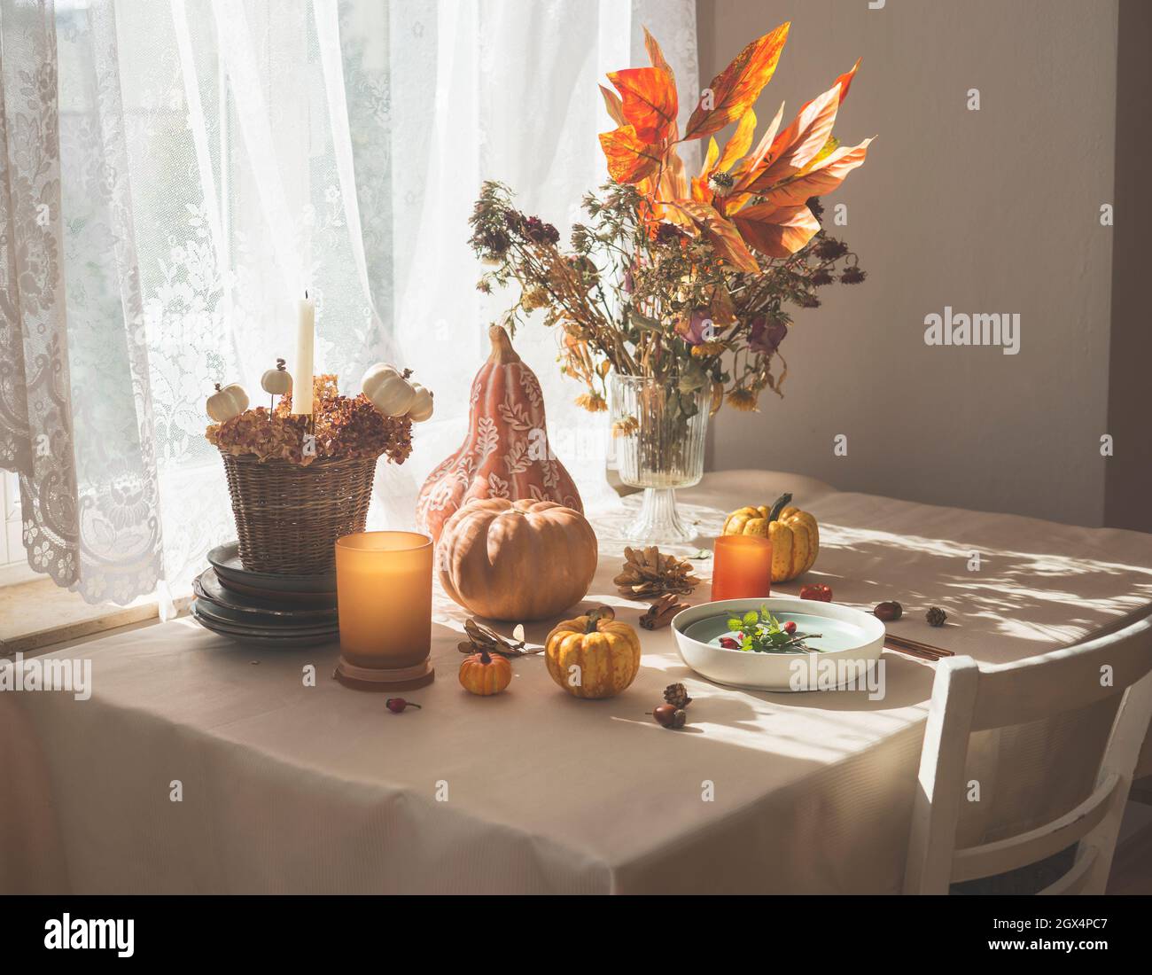 Autumnal decorated kitchen table with various pumpkins, candles, flowers and fall leaves arrangement , plate and cutlery. Morning sunlight from window Stock Photo