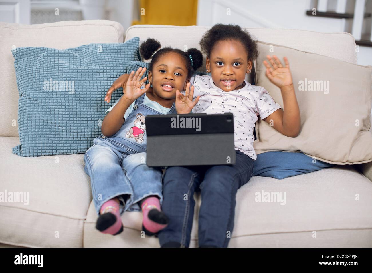 Pretty african american girls smiling and waving hands on camera sitting on cozy sofa with digital tablet. Concept of childhood and technology. Stock Photo