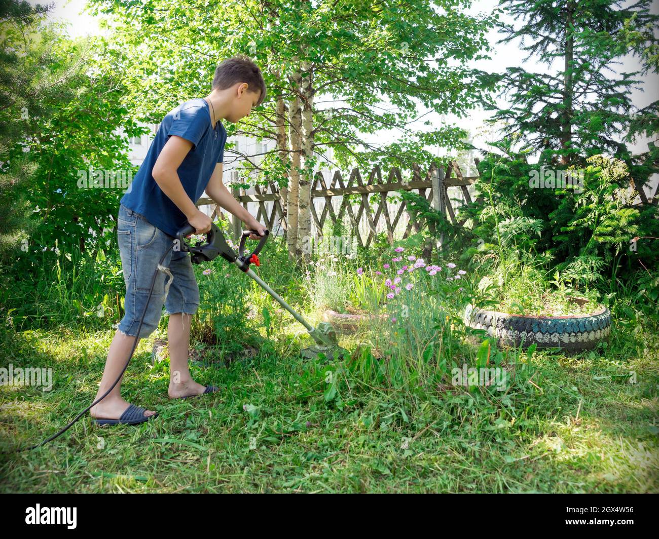 A boy, 11 years old, mows the lawn with an electric scythe near the trees in the yard of a house on a sunny summer day. Stock Photo