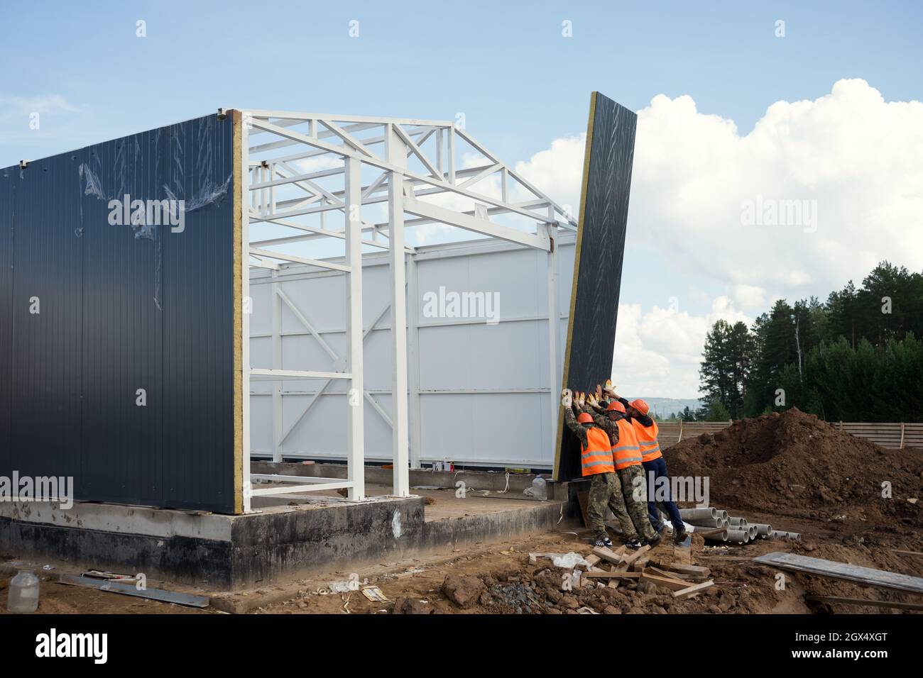Workers install a wall panel with their hands on the frame of an industrial building at a construction site in summer. Stock Photo