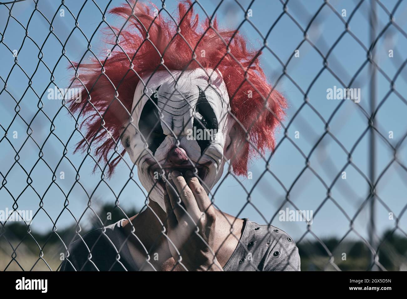 closeup of a scary evil clown, with his hand in his mouth, staring at the observer through a chain-link fence outdoors Stock Photo
