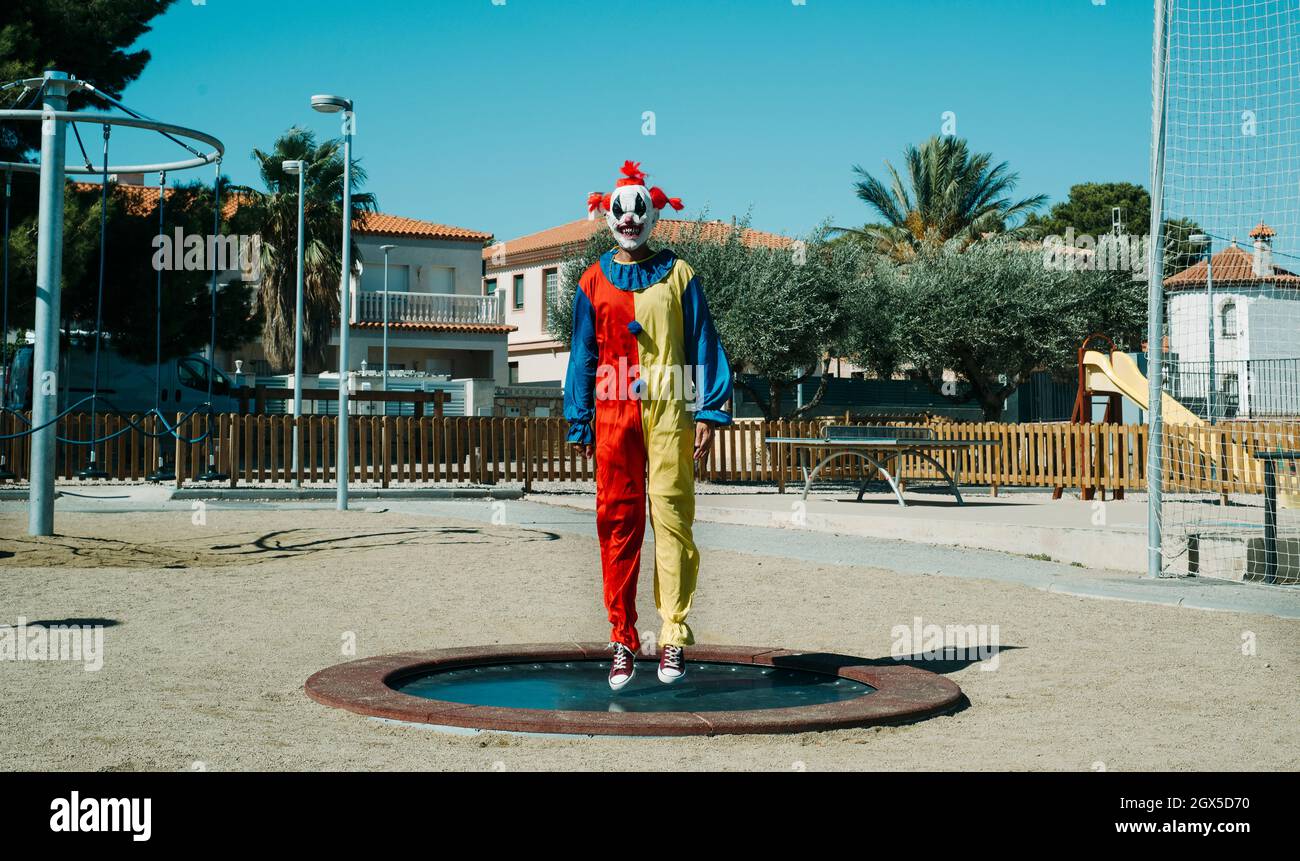 a creepy clown, wearing a colorful yellow, red and blue costume, is bouncing on a trampoline in an outdoor public playground Stock Photo