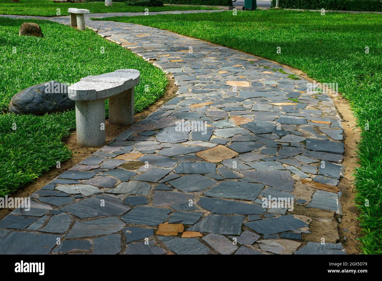 Walkway in the garden paved of stone winds among the green grass. Rest bench in the city park Stock Photo