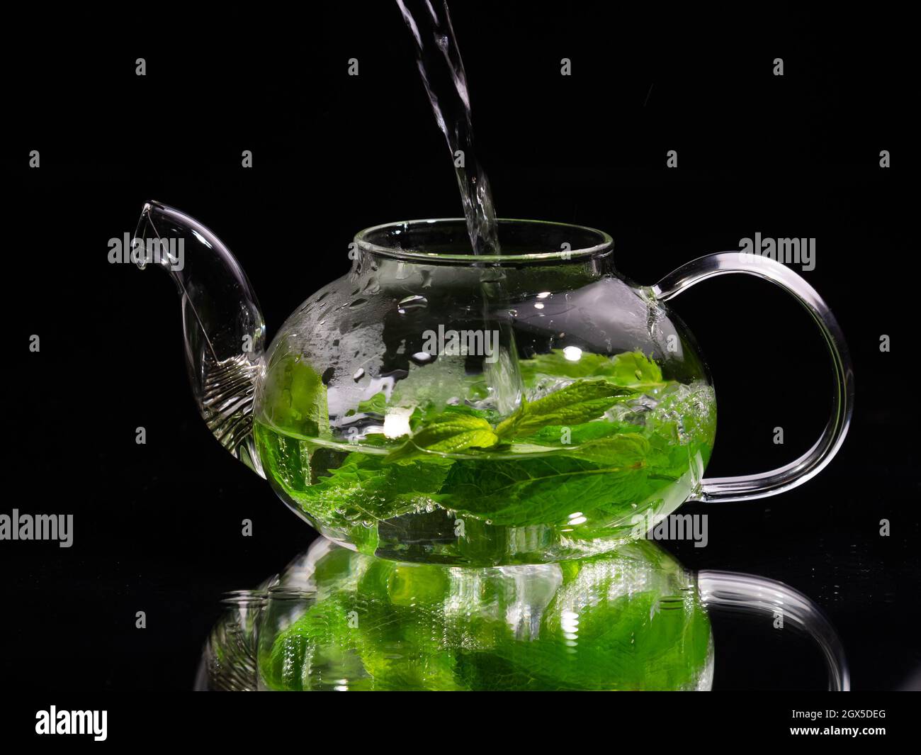 Pouring hot water into a glass teapot on a black background, green mint