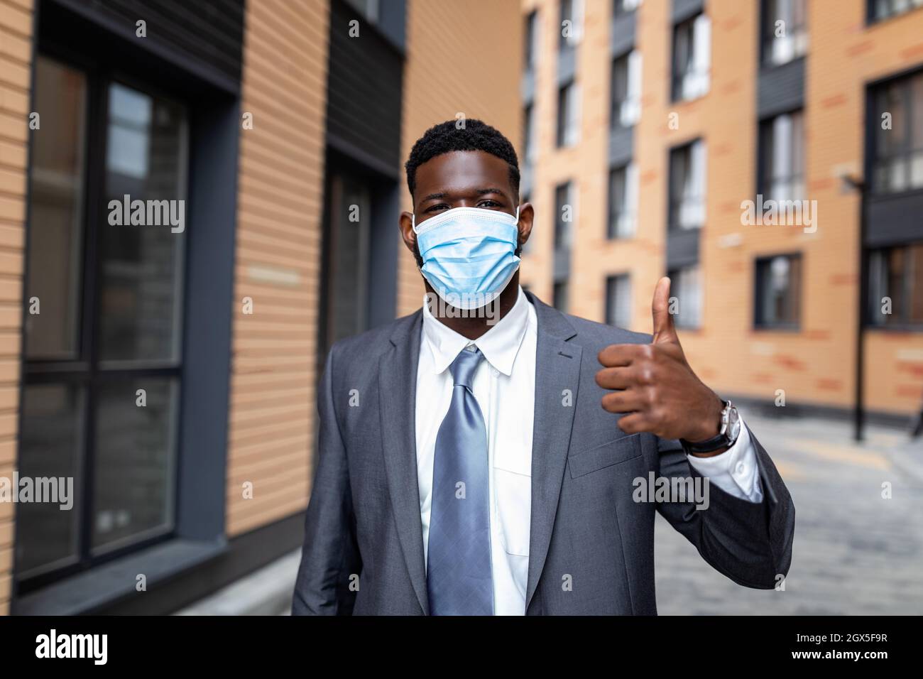 African american businessman in medical face mask and formal outfit, showing thumb up while standing outdoors Stock Photo