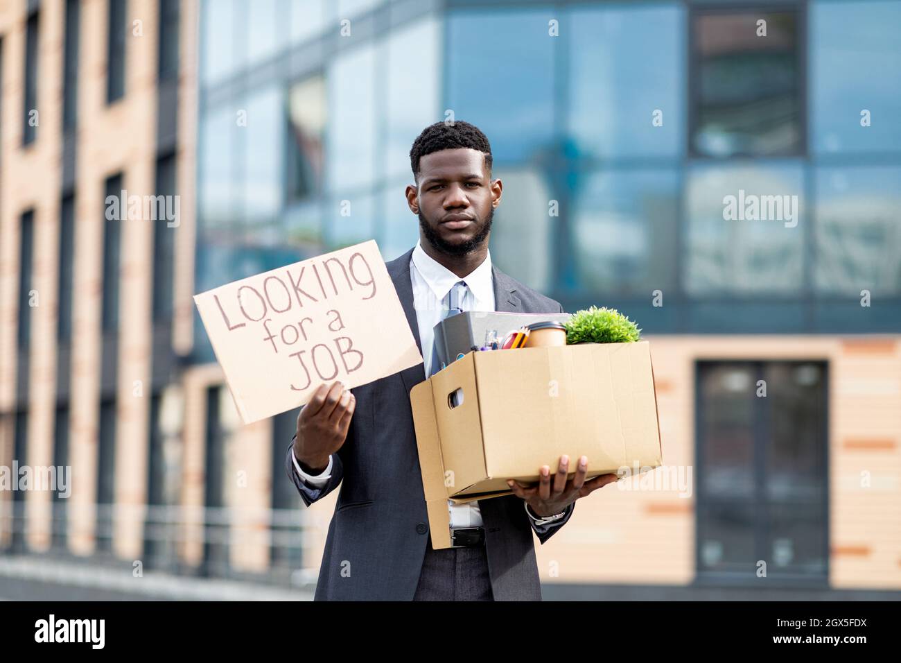 Looking for a job. Upset black businessman with box of personal stuff and poster, got fired, standing outdoors Stock Photo