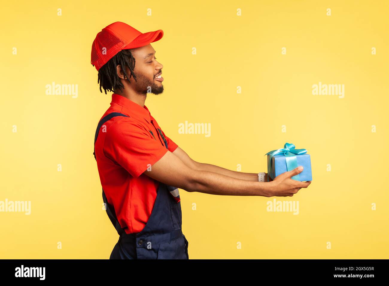 Side view of happy courier in blue overalls and red T-shirt holding wrapped box, smiling friendly to client, carrying birthday present. Indoor studio shot isolated on yellow background. Stock Photo