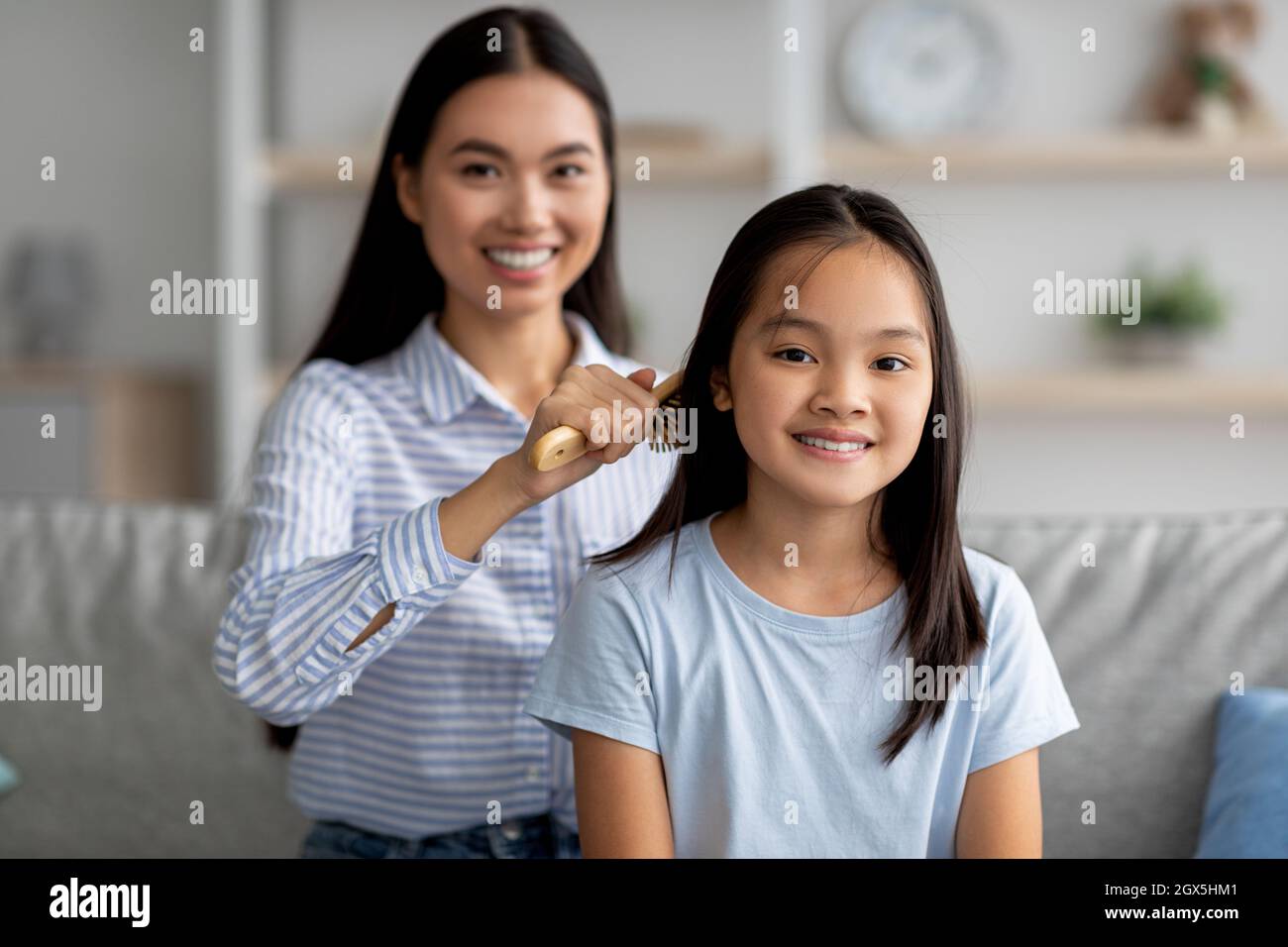 Loving asian mother combing her daughter hair with wooden brush while sitting together on sofa at home Stock Photo