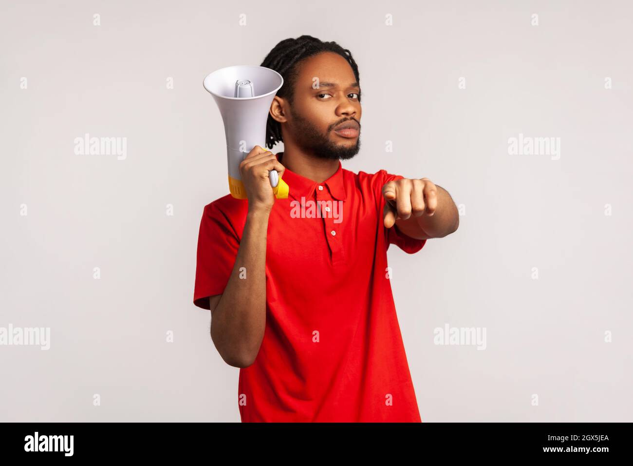 Strict serious man with dreadlocks wearing red casual style T-shirt, pointing finger at camera holding loudspeaker in hand, talking to you, protesting. Indoor studio shot isolated on gray background. Stock Photo