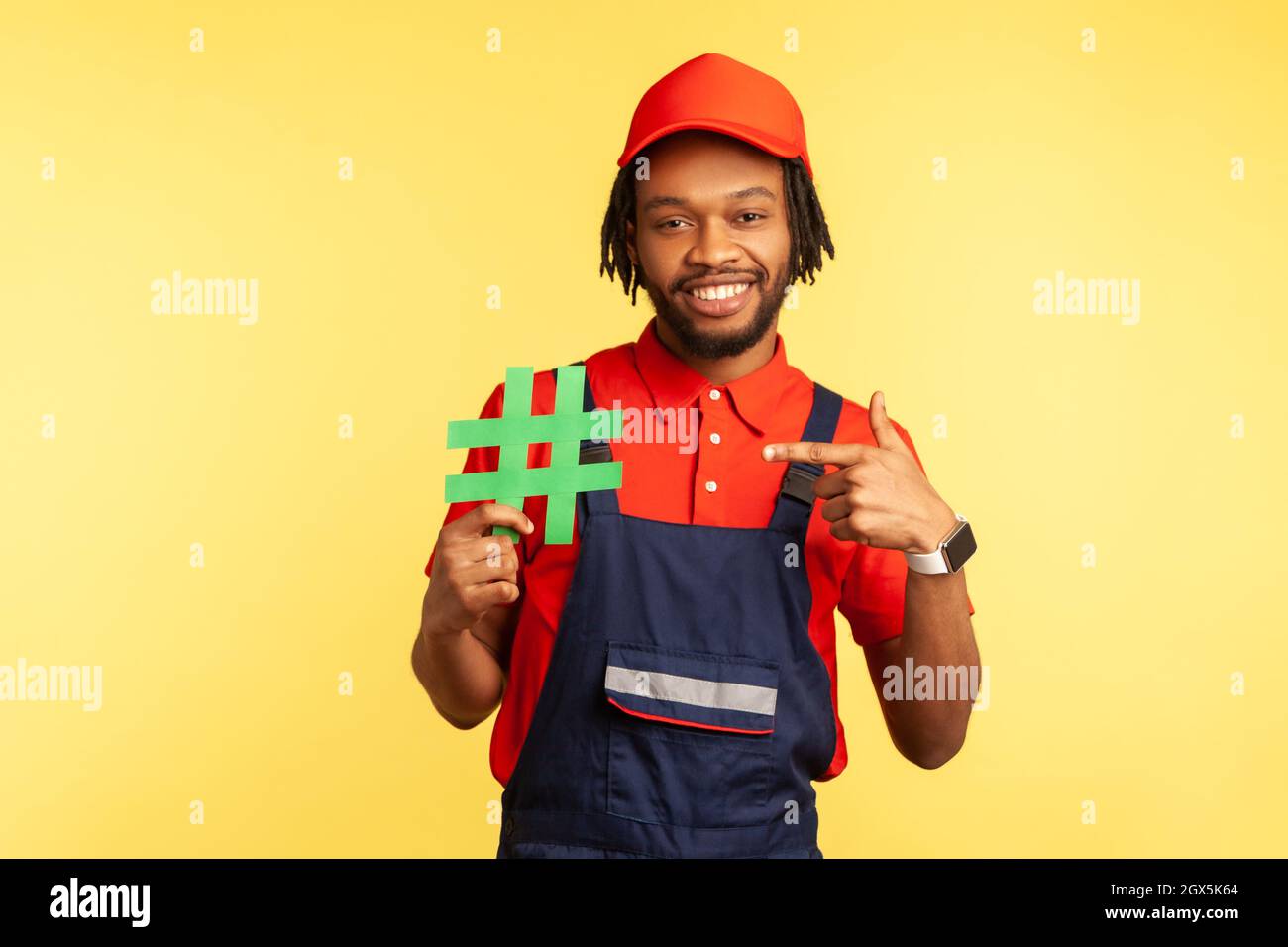 Portrait of happy workman wearing blue overalls standing, holding green hashtag and pointing finger to it, looking at camera with smile. Indoor studio shot isolated on yellow background. Stock Photo