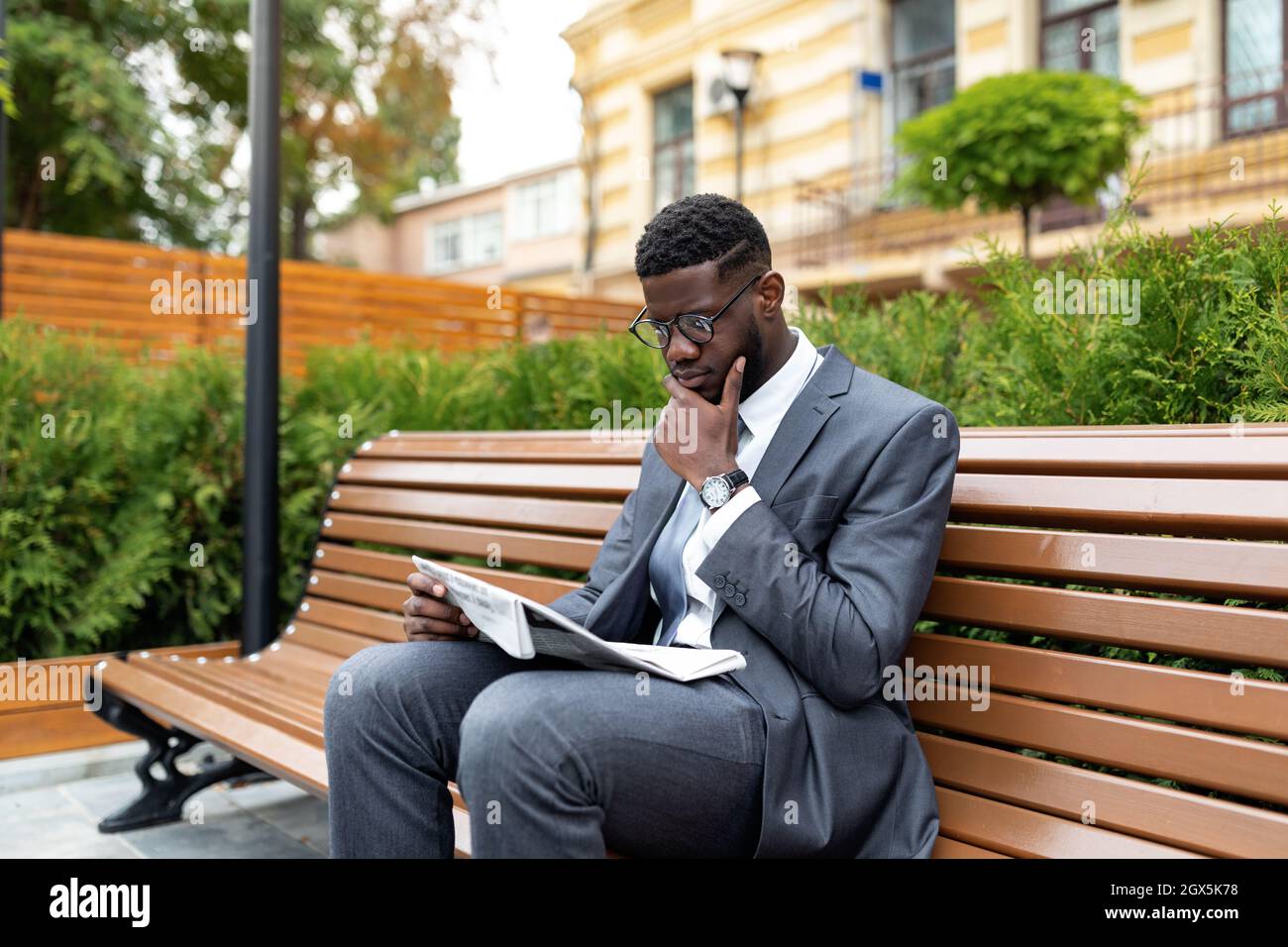 African american businessman in suit reading newspaper while sitting on bench near office center, free space Stock Photo