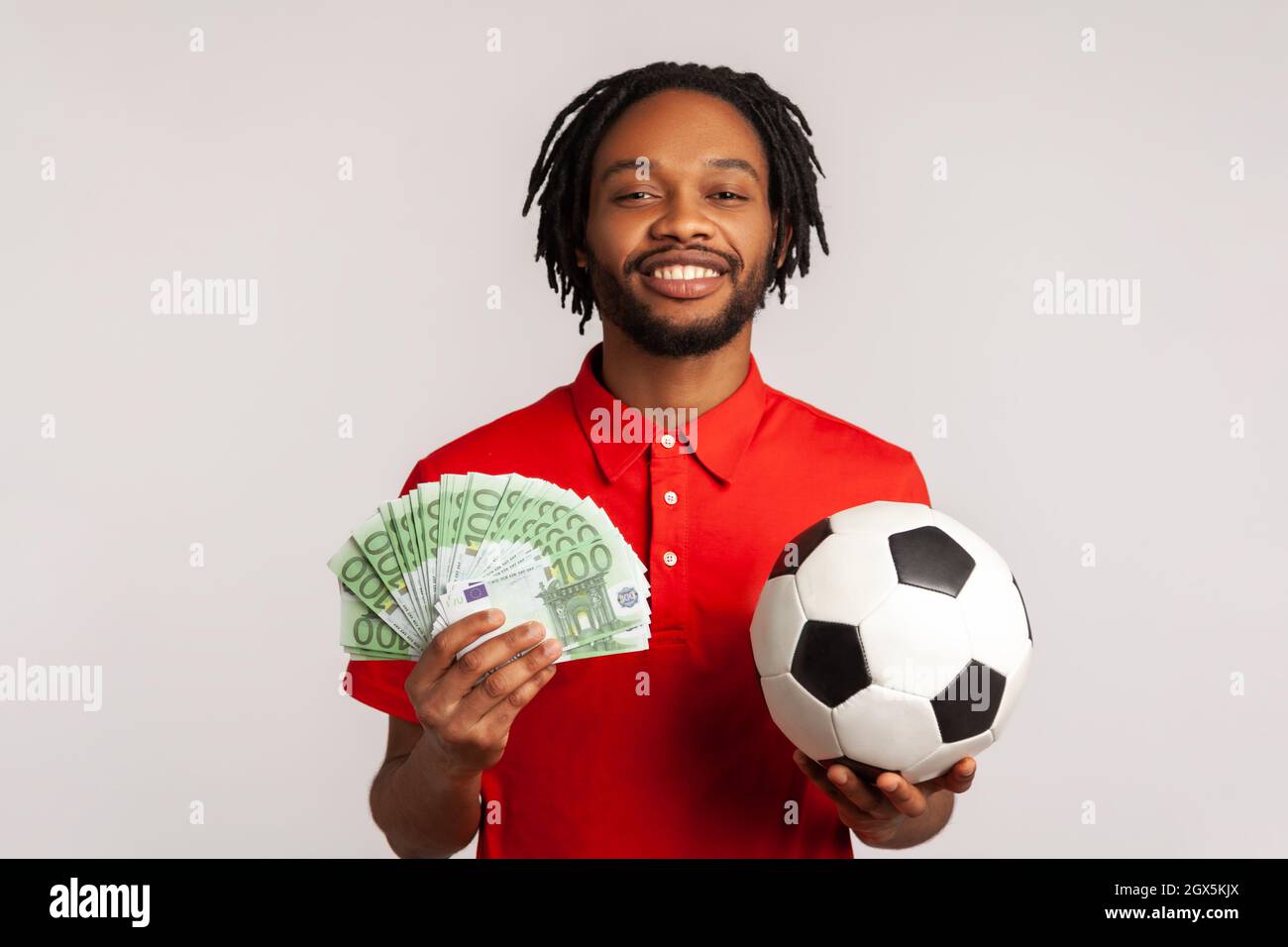 Attractive guy with toothy smile wearing red casual style T-shirt, holding soccer ball and euro banknotes, looking camera, betting and winning. Indoor studio shot isolated on gray background. Stock Photo