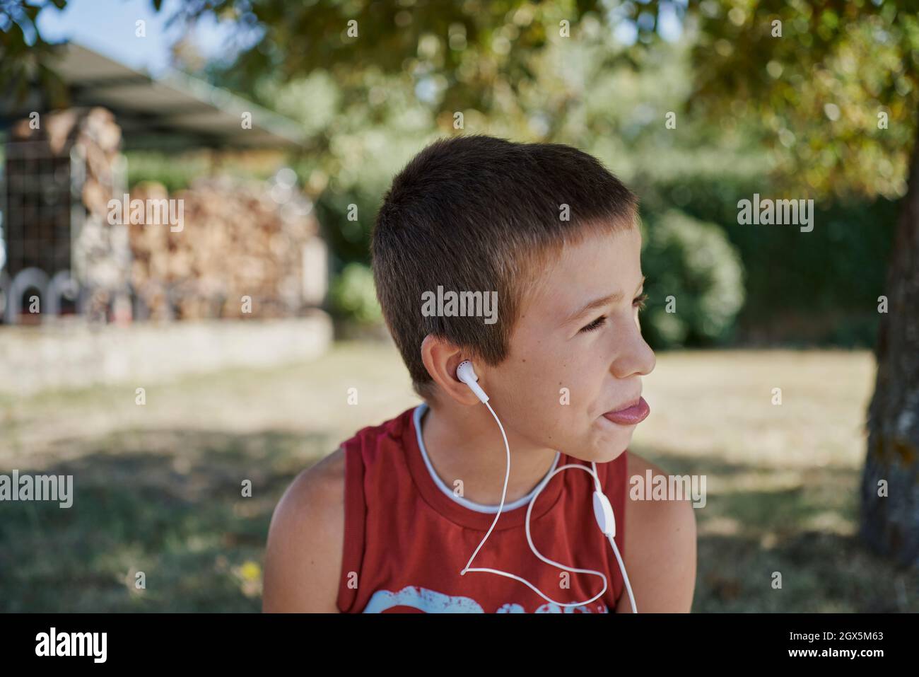 A cute boy listen to music while he looks away under s tree-shade. A Spanish boy uses his white earphones to listen music. Stock Photo