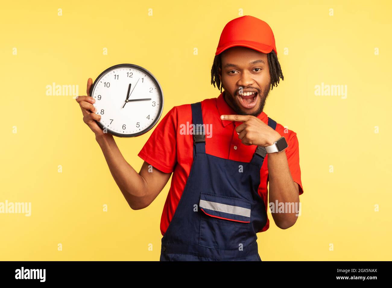 Delivery on time. Optimistic handyman in overalls pointing at clock, smiling to camera, professional and punctual service of house repair, maintenance. Indoor studio shot isolated on yellow background Stock Photo