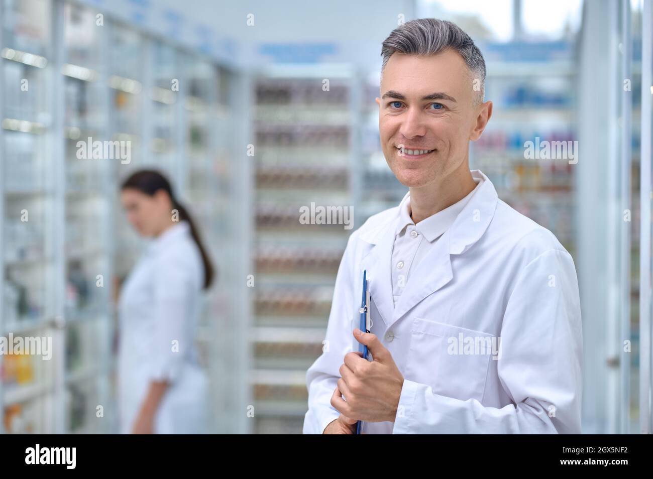 Smiling pharmacist with folder and woman behind Stock Photo