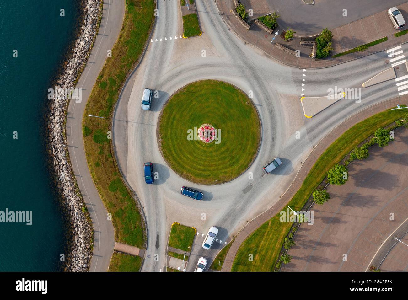 Top view of modern vehicles driving on asphalt roundabout road near calm water in coastal city. Stock Photo