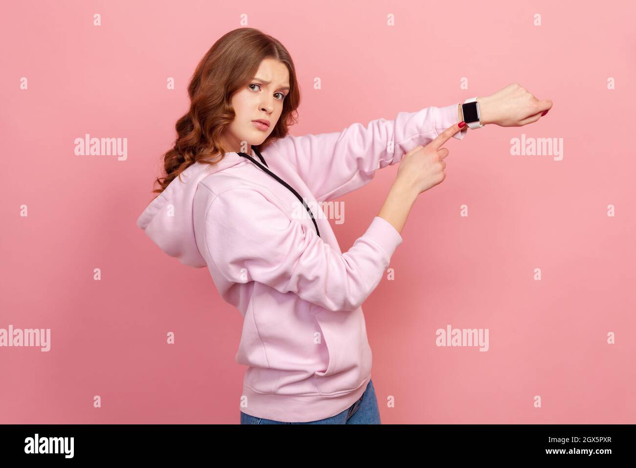 Portrait of displeased young brunette female in hoodie looking impatient and pointing wristwatch, warning of deadline, late hour. Indoor studio shot isolated on pink background Stock Photo