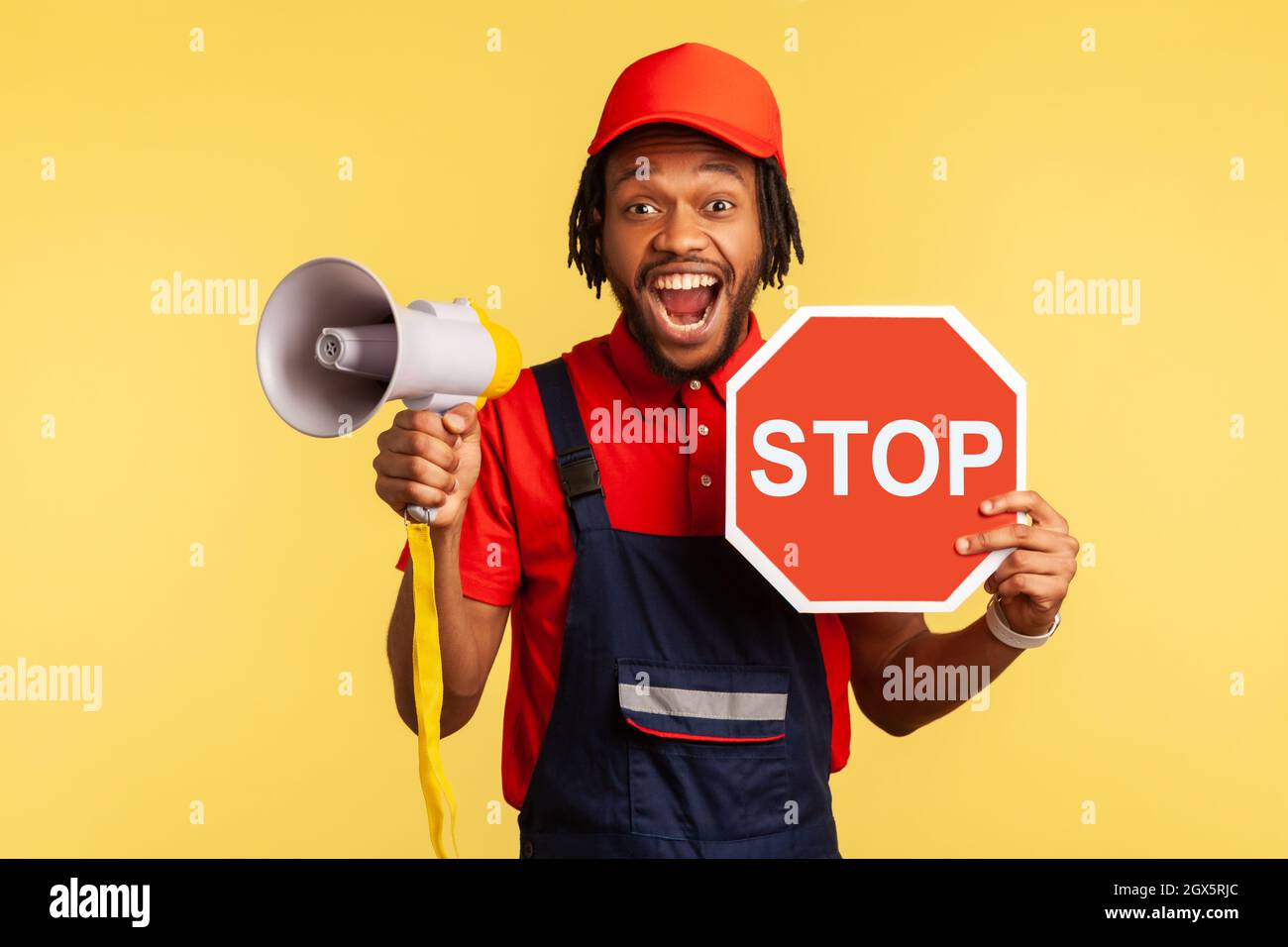 Portrait of excited bearded handyman wearing blue overalls and red T-shirt looking at camera, holding red stop sign and megaphone in his hands. Indoor studio shot isolated on yellow background. Stock Photo