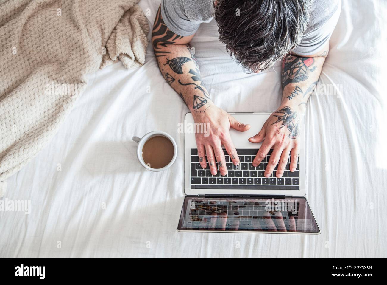cropped view of tattooed man typing on laptop in bed with coffee cup and laptops Stock Photo