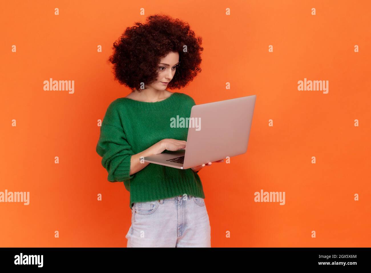 Portrait of attractive concentrated woman with Afro hairstyle wearing green casual style sweater holding laptop, freelancer working online. Indoor studio shot isolated on orange background. Stock Photo