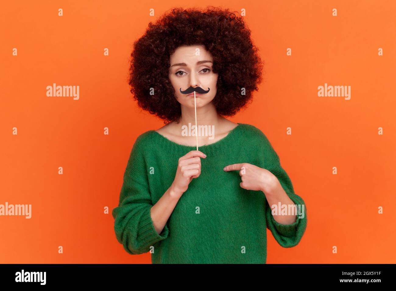 Portrait of funny serious woman with Afro hairstyle wearing green casual style sweater pointing at herself and holding paper mustache. Indoor studio shot isolated on orange background. Stock Photo