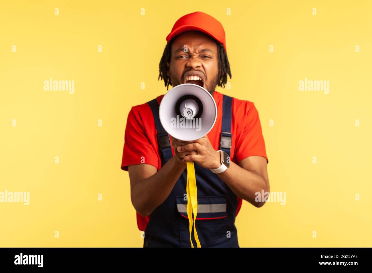 Portrait of angry bearded worker wearing red T-shirt and blue overalls holding megaphone and screaming with aggressive expression, protesting. Indoor studio shot isolated on yellow background. Stock Photo
