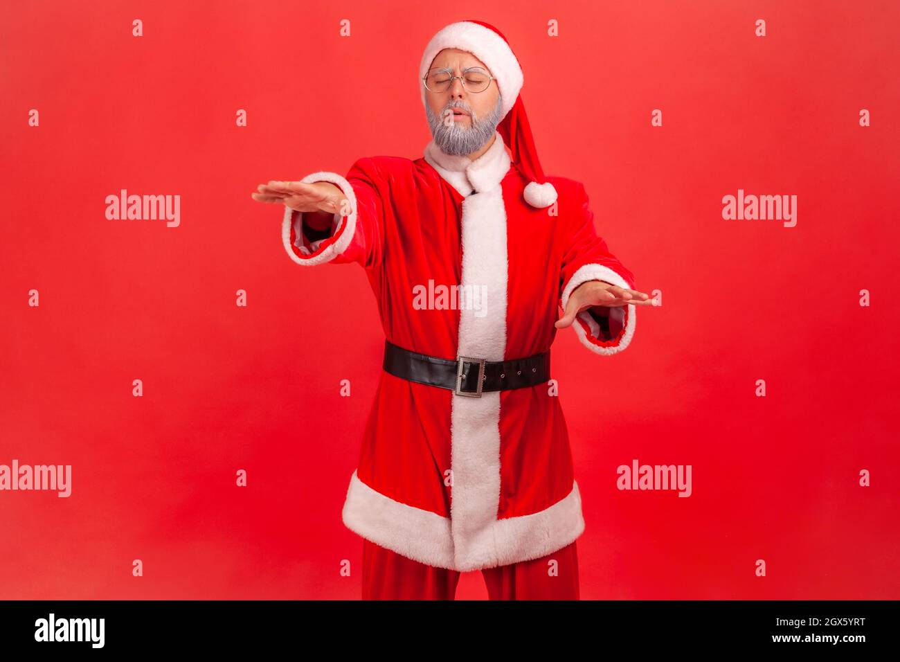 Vision problems. Portrait of blind disoriented lonely elderly man in santa claus costume walking with closed eyes, raising hands to search lost road. Indoor studio shot isolated on red background. Stock Photo