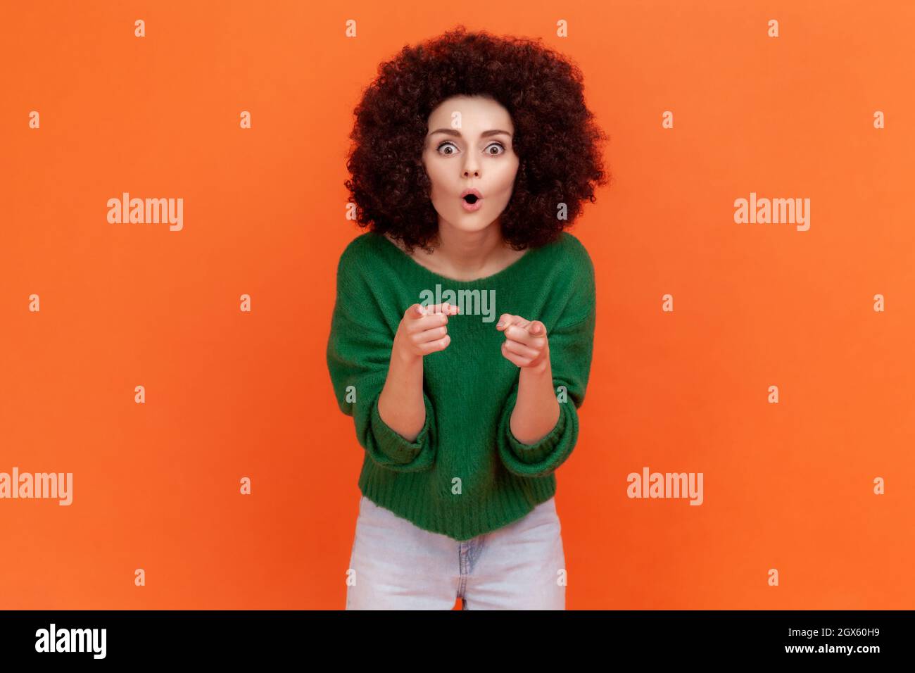 Shocked woman with Afro hairstyle wearing green casual style sweater with open mouth pointing to you, making choice and expressing astonishment. Indoor studio shot isolated on orange background. Stock Photo