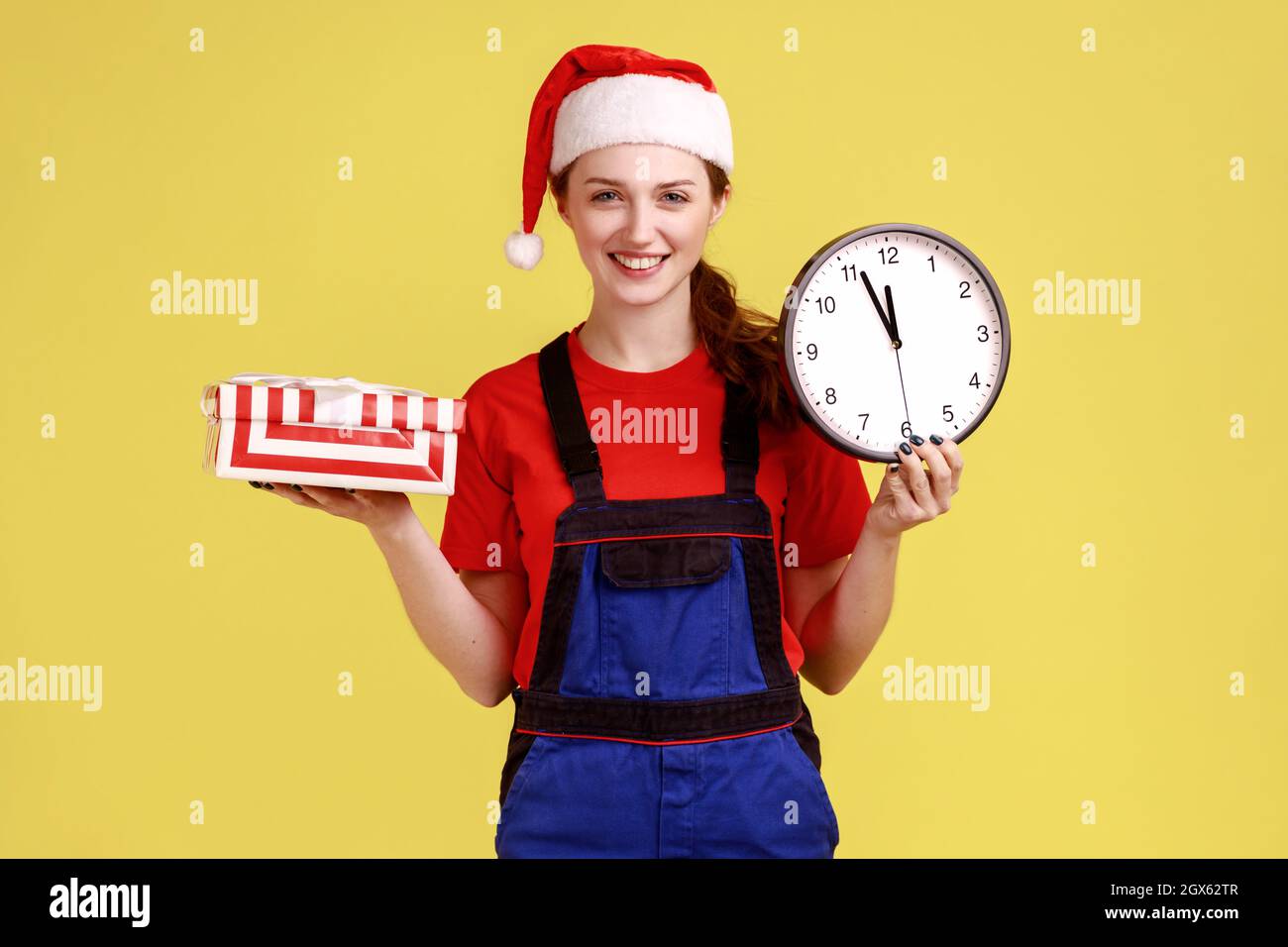 Smiling courier woman holding wrapped present box and wall clock, fast delivery service in time, wearing blue overalls and santa claus hat. Indoor studio shot isolated on yellow background. Stock Photo