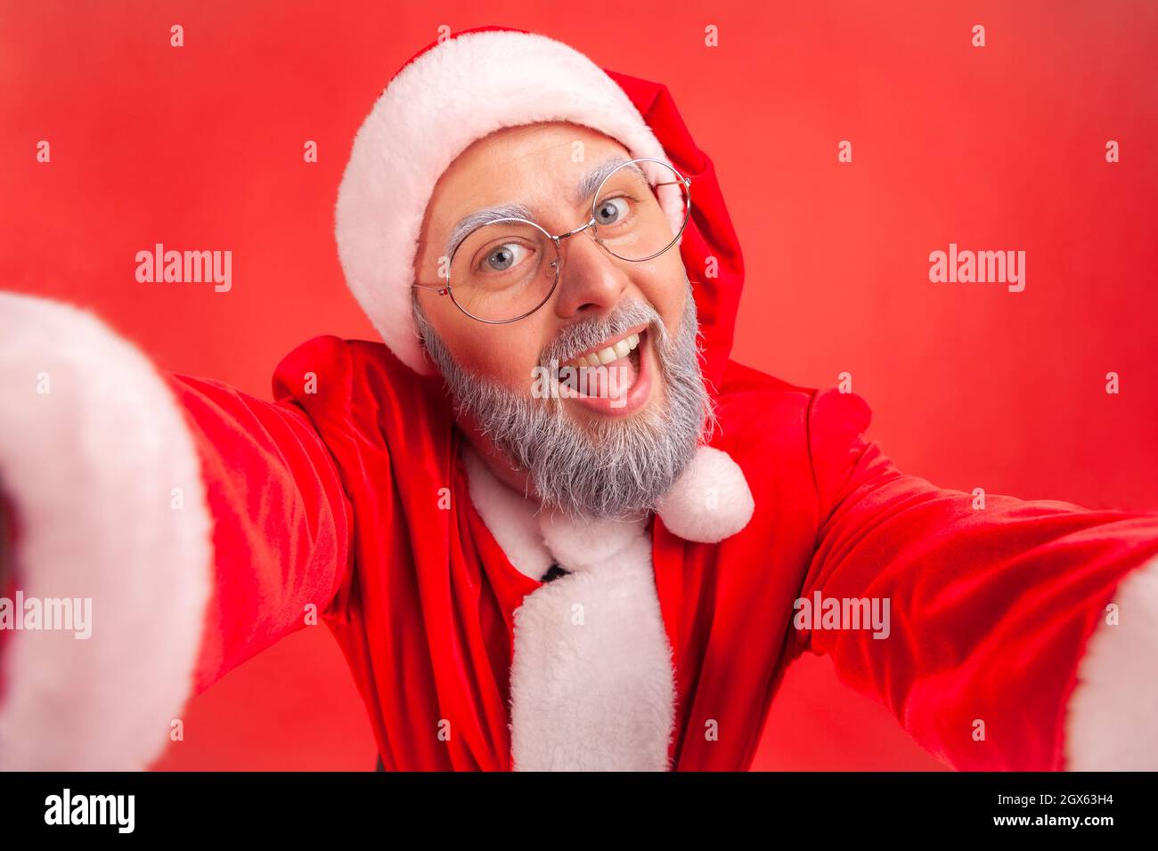Happy funny elderly man with gray beard wearing santa claus costume making selfie POV, looking at camera with smile and positive expression. Indoor studio shot isolated on red background. Stock Photo