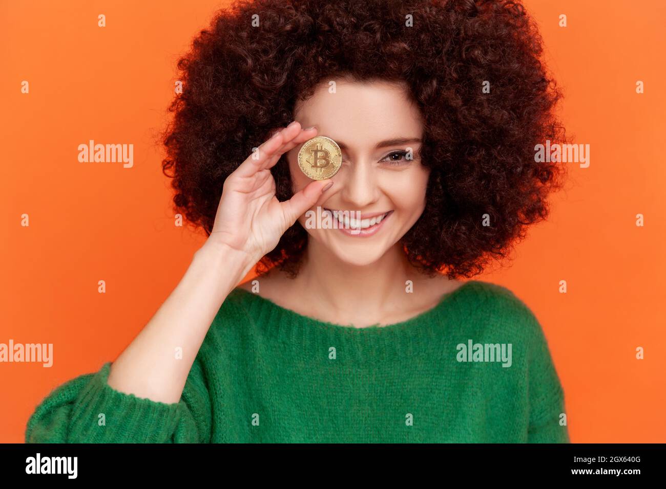 Portrait of happy smiling woman with Afro hairstyle wearing green casual style sweater covering eye with gold bitcoin, blockchain concept. Indoor studio shot isolated on orange background. Stock Photo