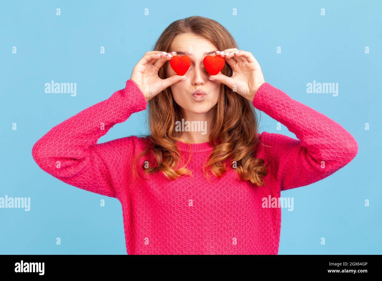 Portrait of romantic woman wearing pink pullover, covering eyes with little toy hearts as if looking with love, affection, sending air kisses. Indoor studio shot isolated on blue background. Stock Photo