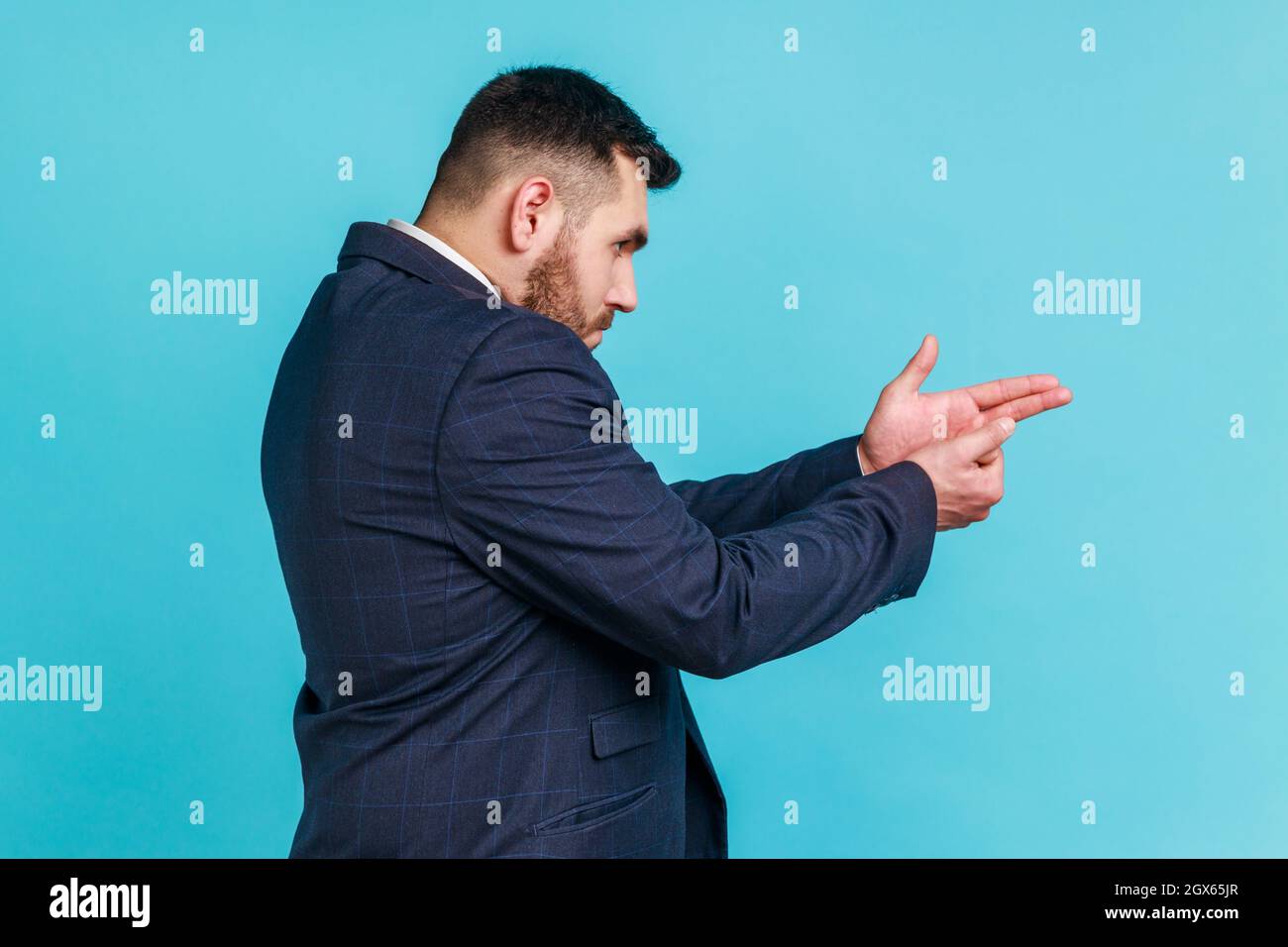 Side view of dangerous businessman wearing official style suit pointing finger gun, threatening to kill, aiming hand pistol, forward choosing direction. Indoor studio shot isolated on blue background. Stock Photo