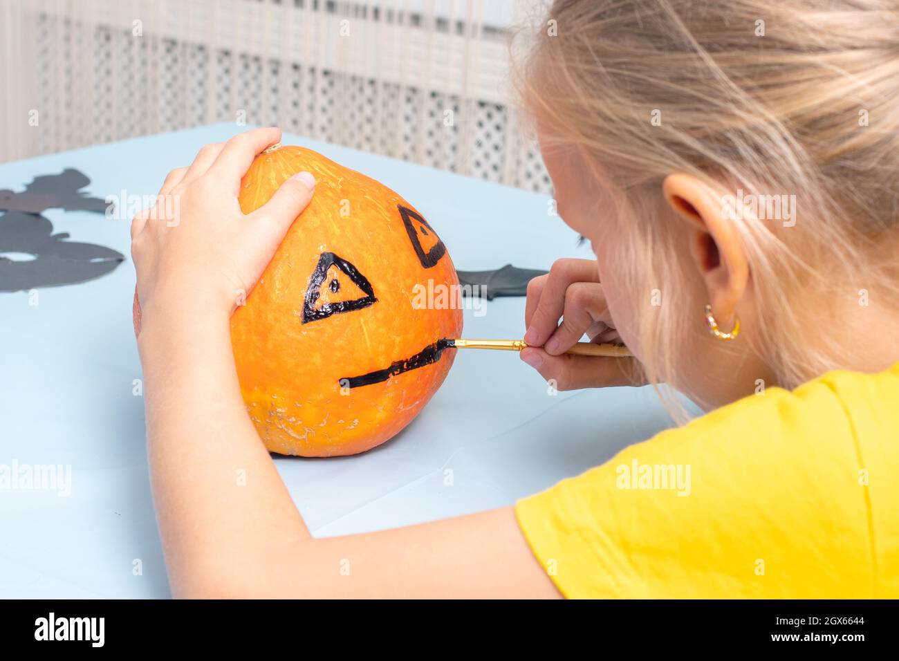 Cute young girl painting scary face on orange pumpkin with brush for home decoration during Halloween holiday, back view, close-up. Halloween party an Stock Photo