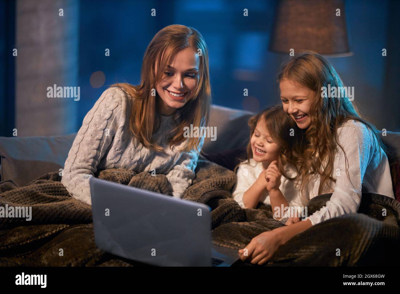 Two little sisters and their mother sitting on couch under soft blanket ...