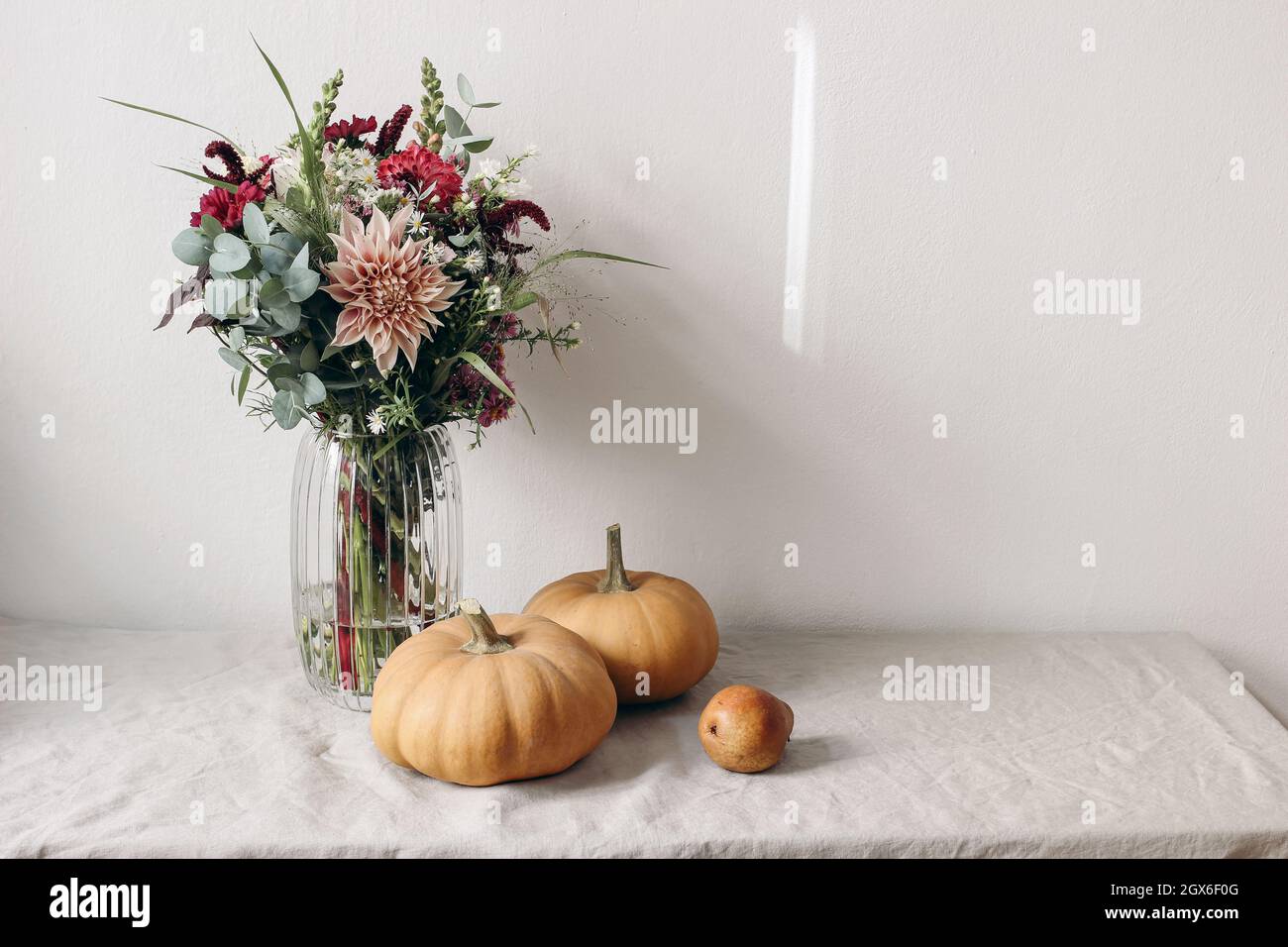 Autumn still life scene. Orange pumkins, pear fruit on linen table cloth in sunlight. Floral bouquet of dahlia flowers, cosmos and eucalyptus in glass Stock Photo