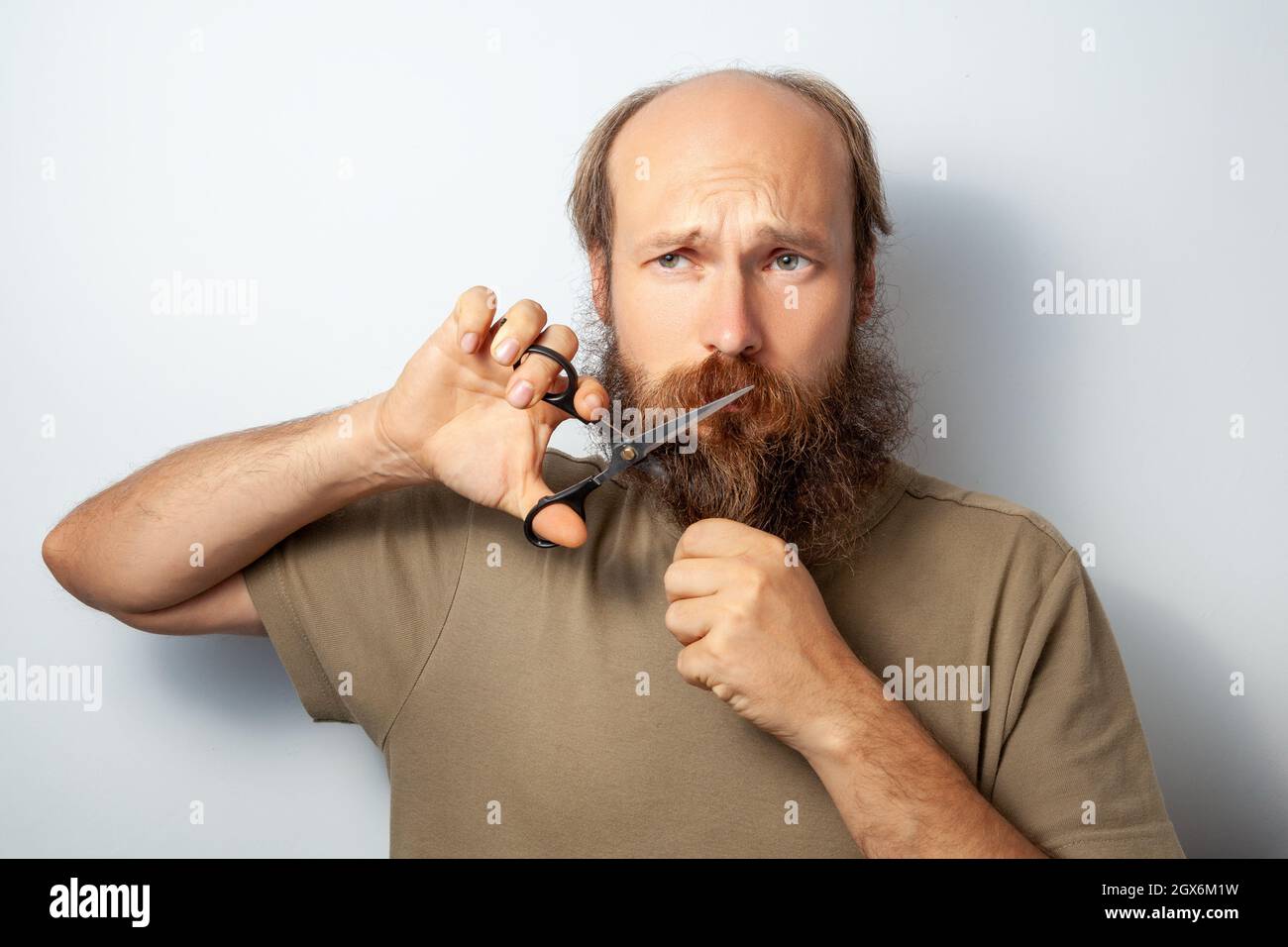 Adult handsome male cutting of his beard with scissors, looking away with regret in his eyes, bald bearded man wearing casual style T-shirt. Indoor studio shot isolated on gray background. Stock Photo