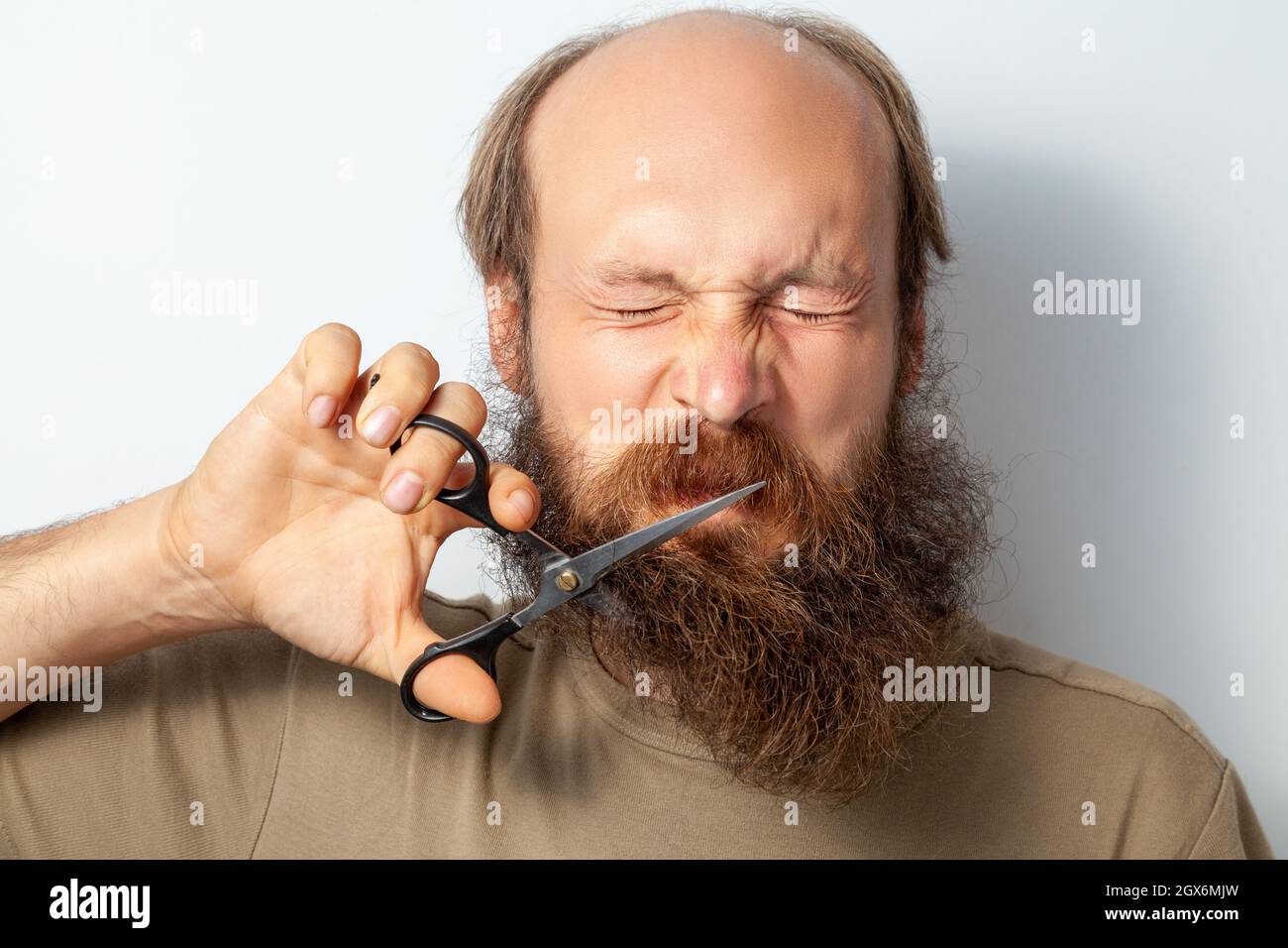 Portrait of male cutting of his beard, screws up his eyes, feels regret, holding scissors in hands, bald bearded man wearing T-shirt. Indoor studio shot isolated on gray background. Stock Photo