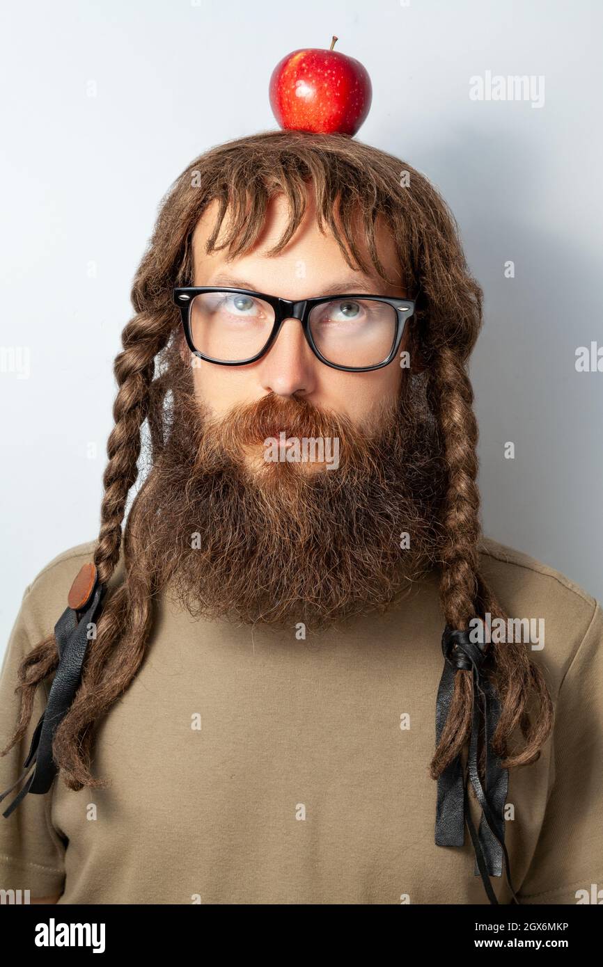 Handsome male scientist or professor with long beard and pigtails holding apple on head, newtons law, bald bearded man wearing T-shirt and glasses. Indoor studio shot isolated on gray background. Stock Photo