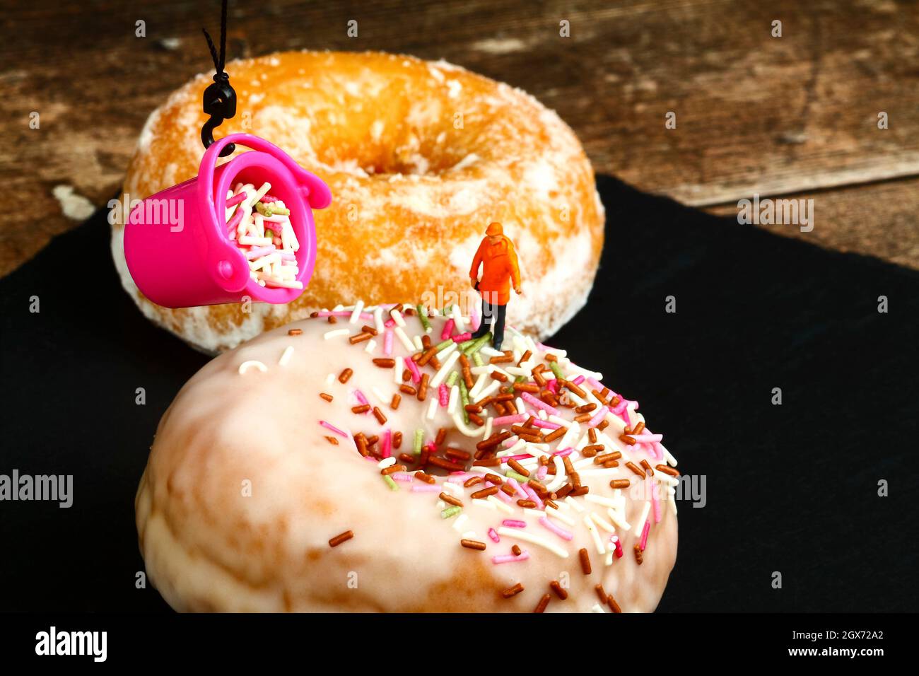 Conceptual image of a miniature figure workman guiding a bucket filled with sprinkles to pour over the icing sugar topping of a fresh doughnut Stock Photo