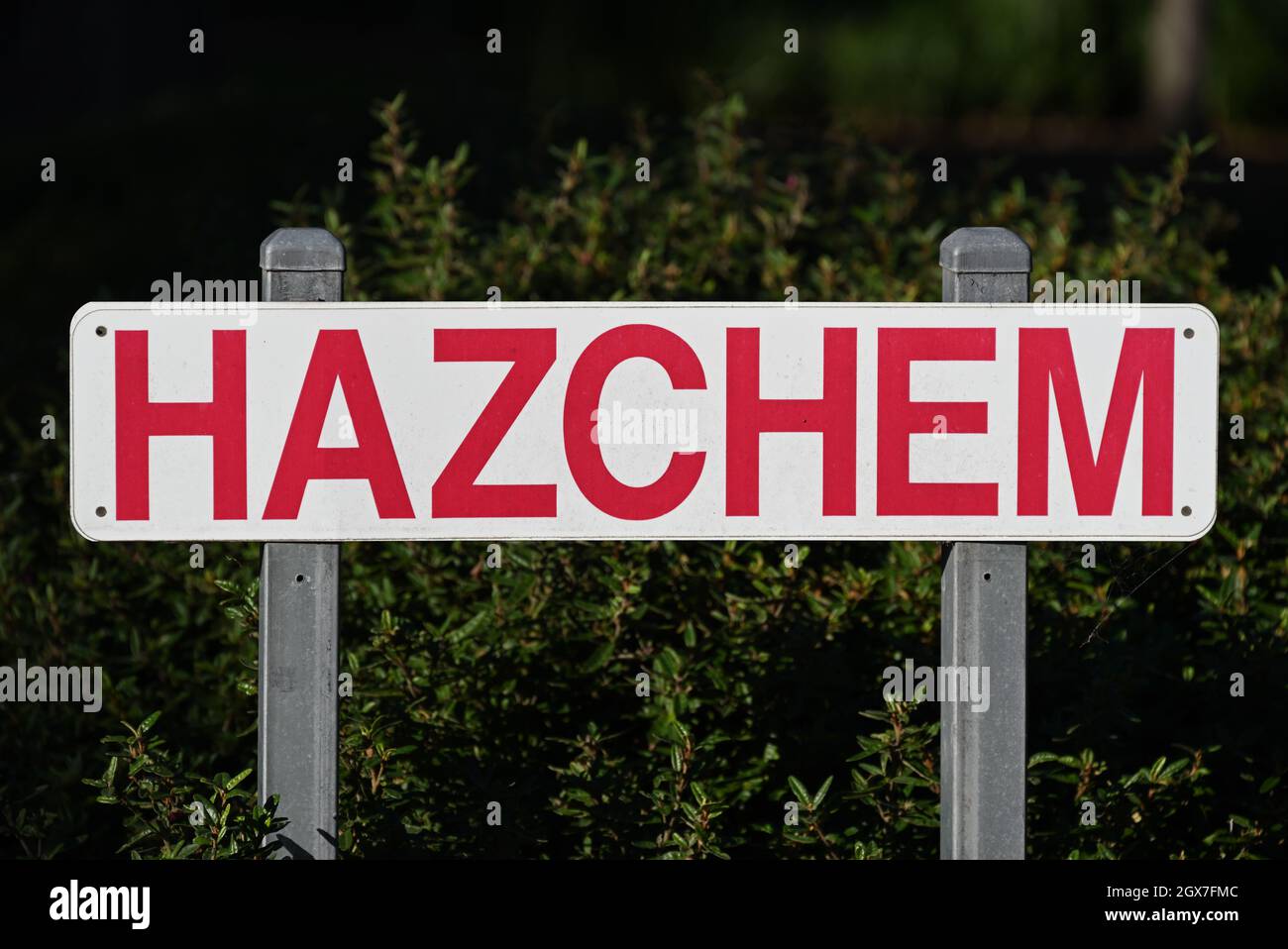 White hazchem sign with bright red lettering, held up by two steel posts. The dirty sign sits in front of a dark green bush, spiderweb hanging off it Stock Photo