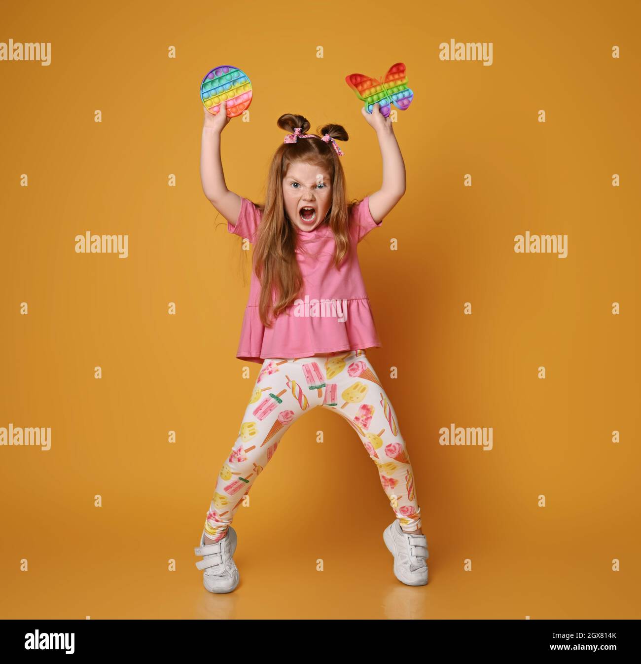 Spoiled, screaming loudly kid girl in colorful clothes going to throw two sensory rainbow color toys - round pop it Stock Photo