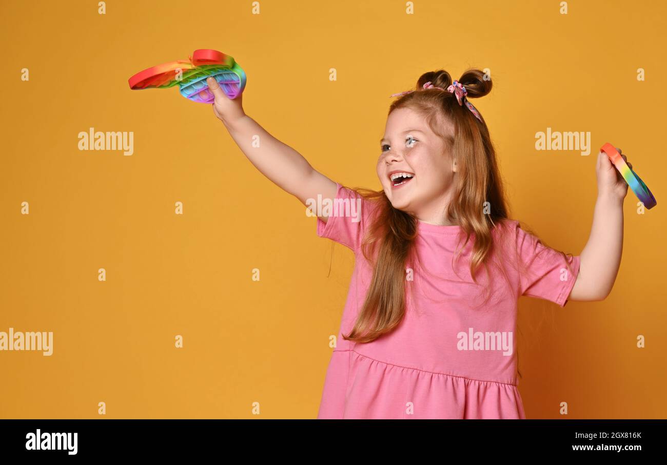 Happy redhead kid girl in pink shirt plays with new sensory rainbow color toys - butterfly shape and round pop it Stock Photo