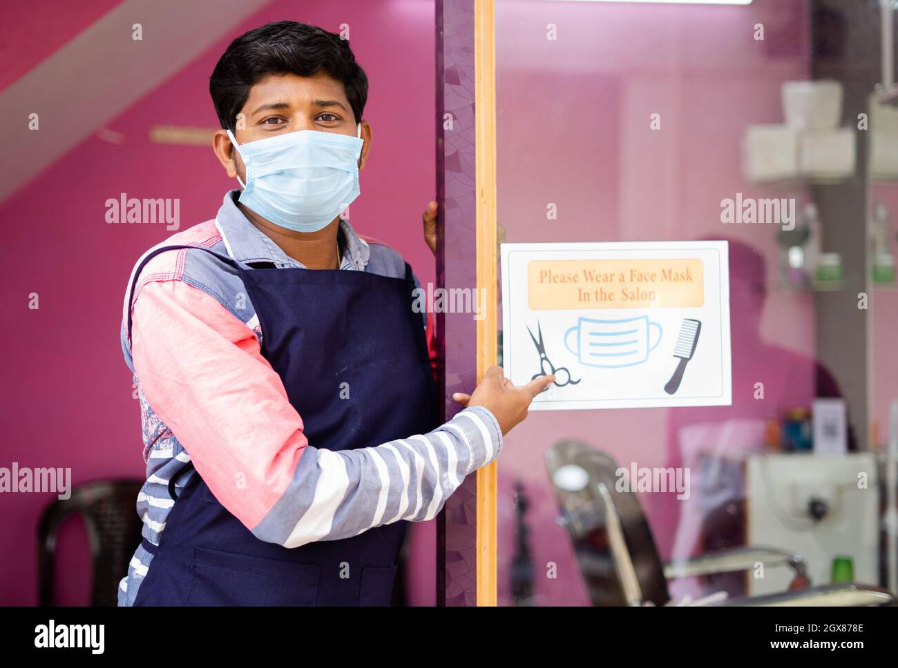 Barber with medical mask showing wear mask at saloon sign board to avoid coronavirus or covid-19 infection as safety measures Stock Photo