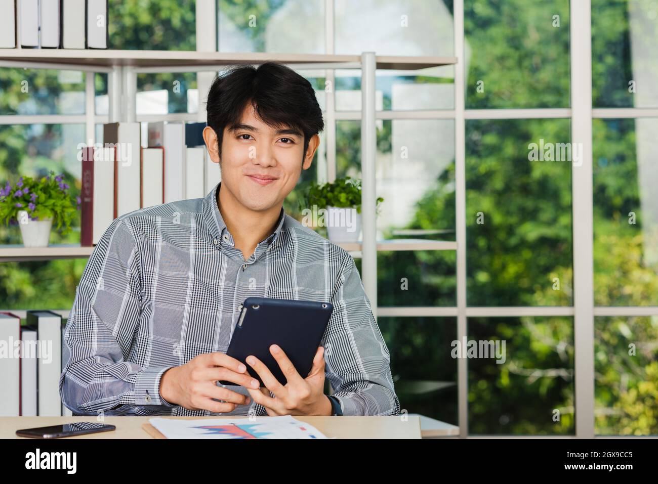Asian young businessman sitting on desk office he smiles and working on a new tablet computer, the confident young handsome man using touchpad checkin Stock Photo
